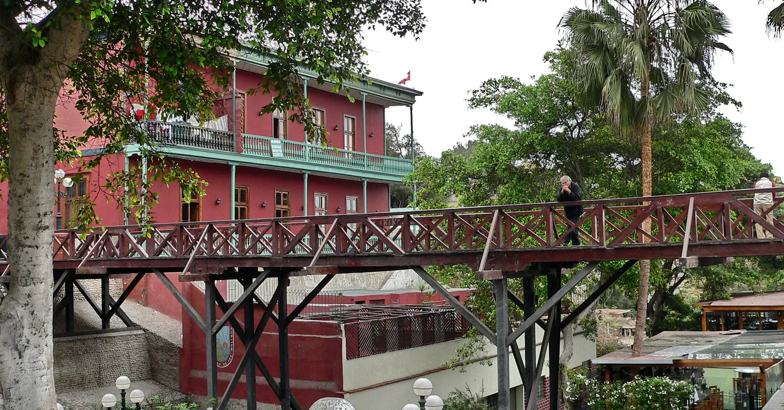 The wooden Puente de los Suspiros seen side-on in Barranco, Lima, its dark timber trestles carrying a lattice-railed deck past a deep red three-storey house with green balconies
