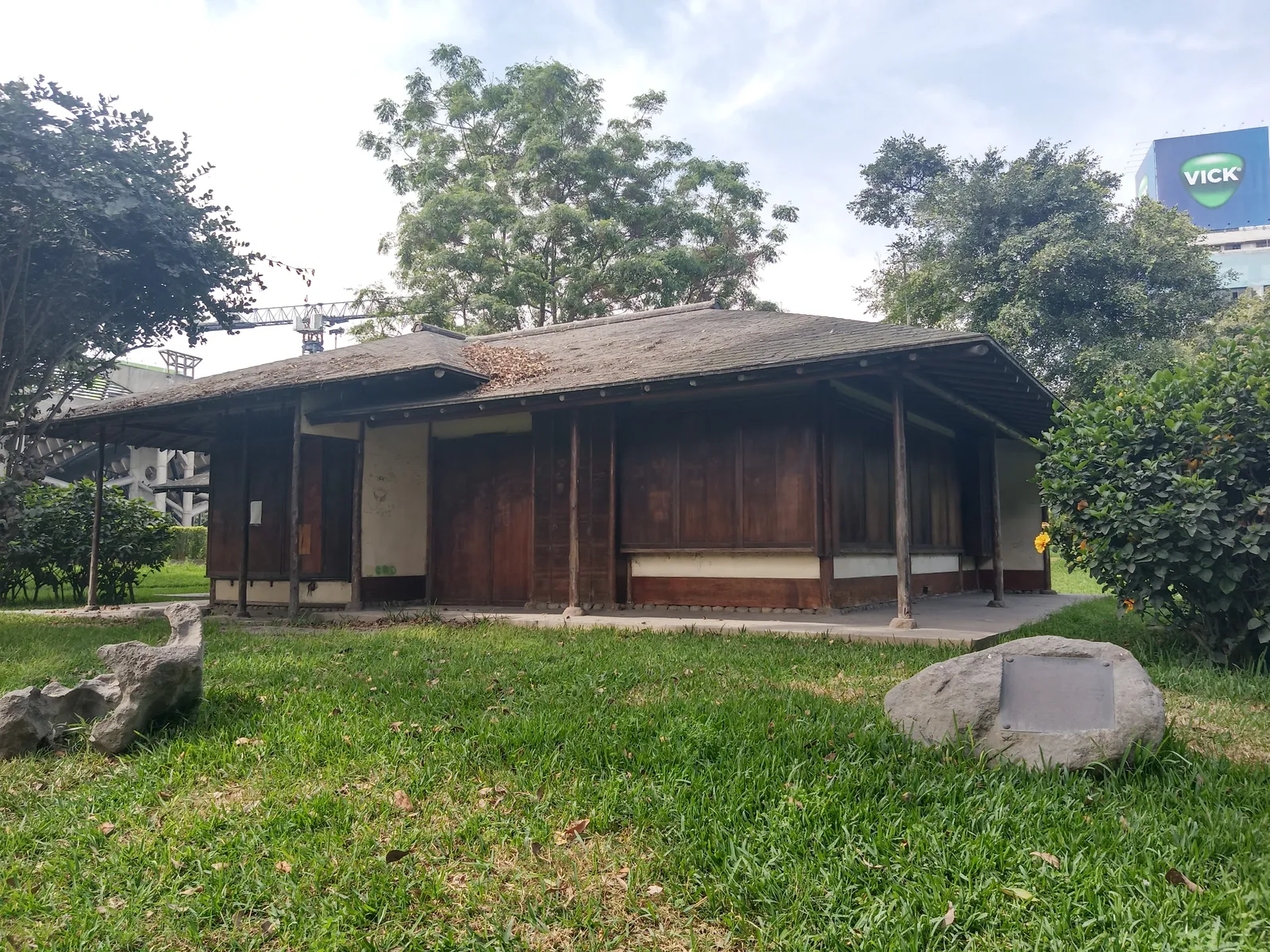 A low wooden pavilion with a wide overhanging roof standing on lawn in the Japanese garden of Lima's Parque de la Exposición, with rocks set into the grass in front of it
