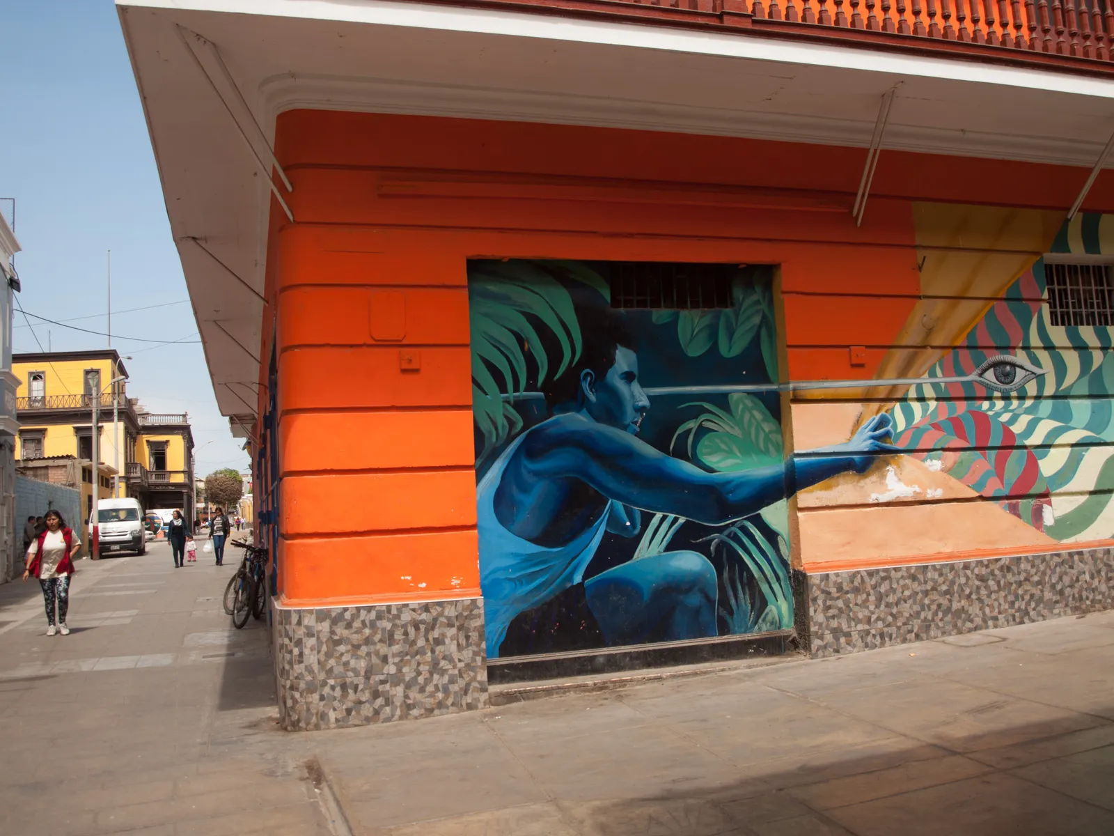 A mural of a figure among green leaves painted across the corner of an orange building in Callao's old port district, with people walking the pavement beyond