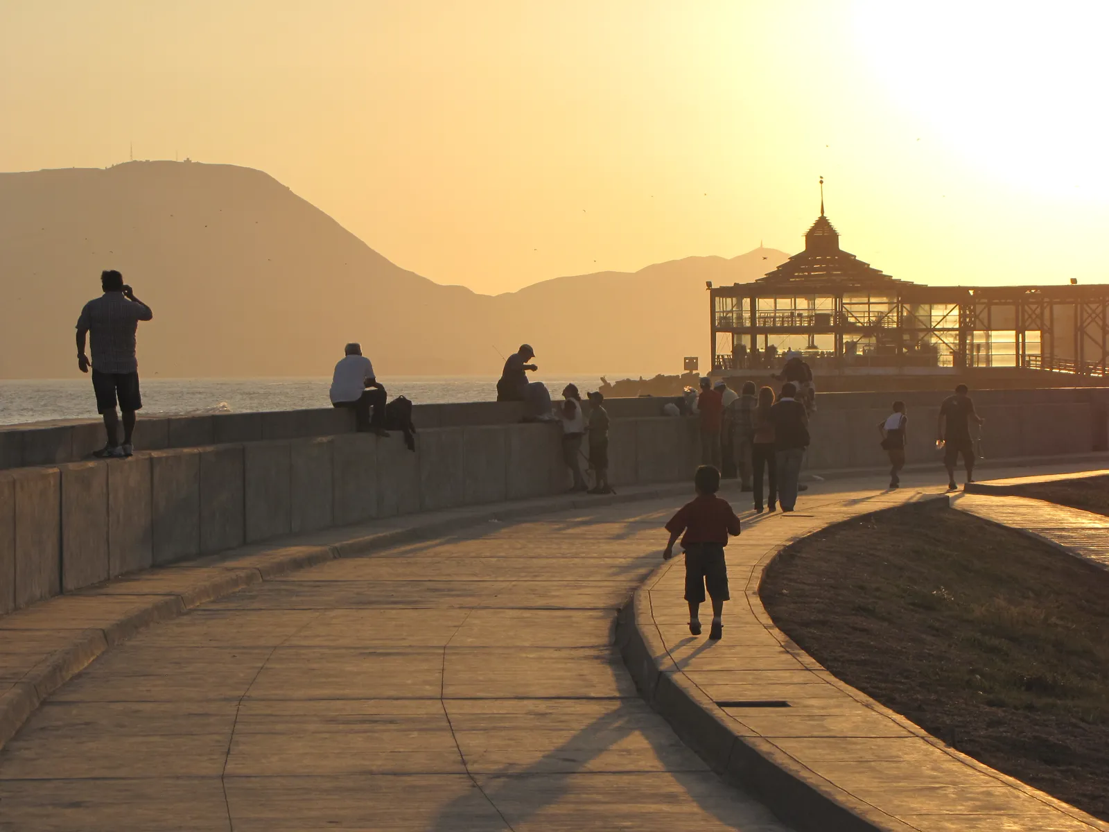 The curving seawall promenade at La Punta, Callao, in low evening light, with people sitting on the wall above the water and a domed pavilion further along