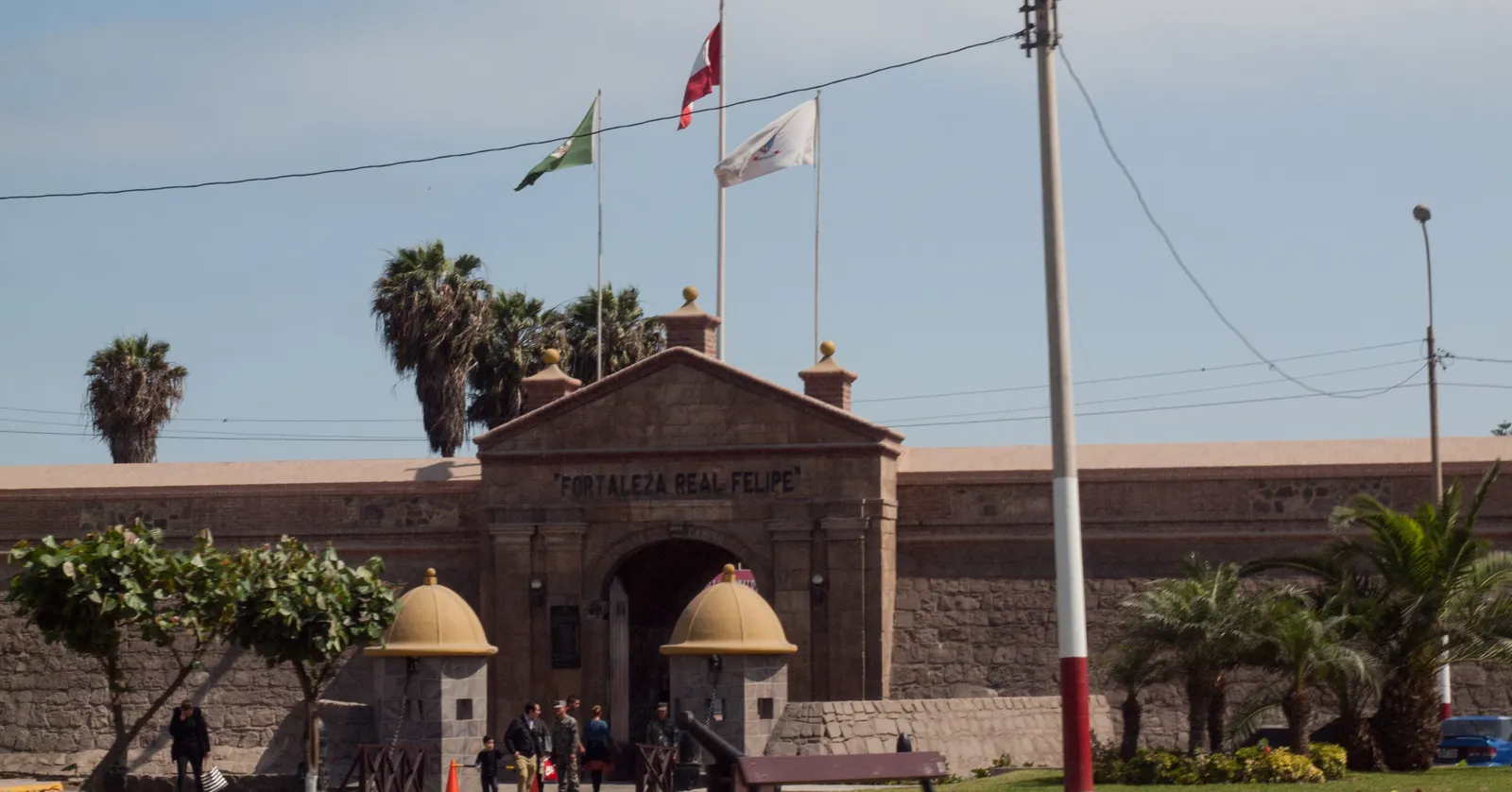 The stone gatehouse of the Real Felipe fortress in Callao, Peru, with its name carved above the arch, three flags flying over the curtain wall and palms behind it