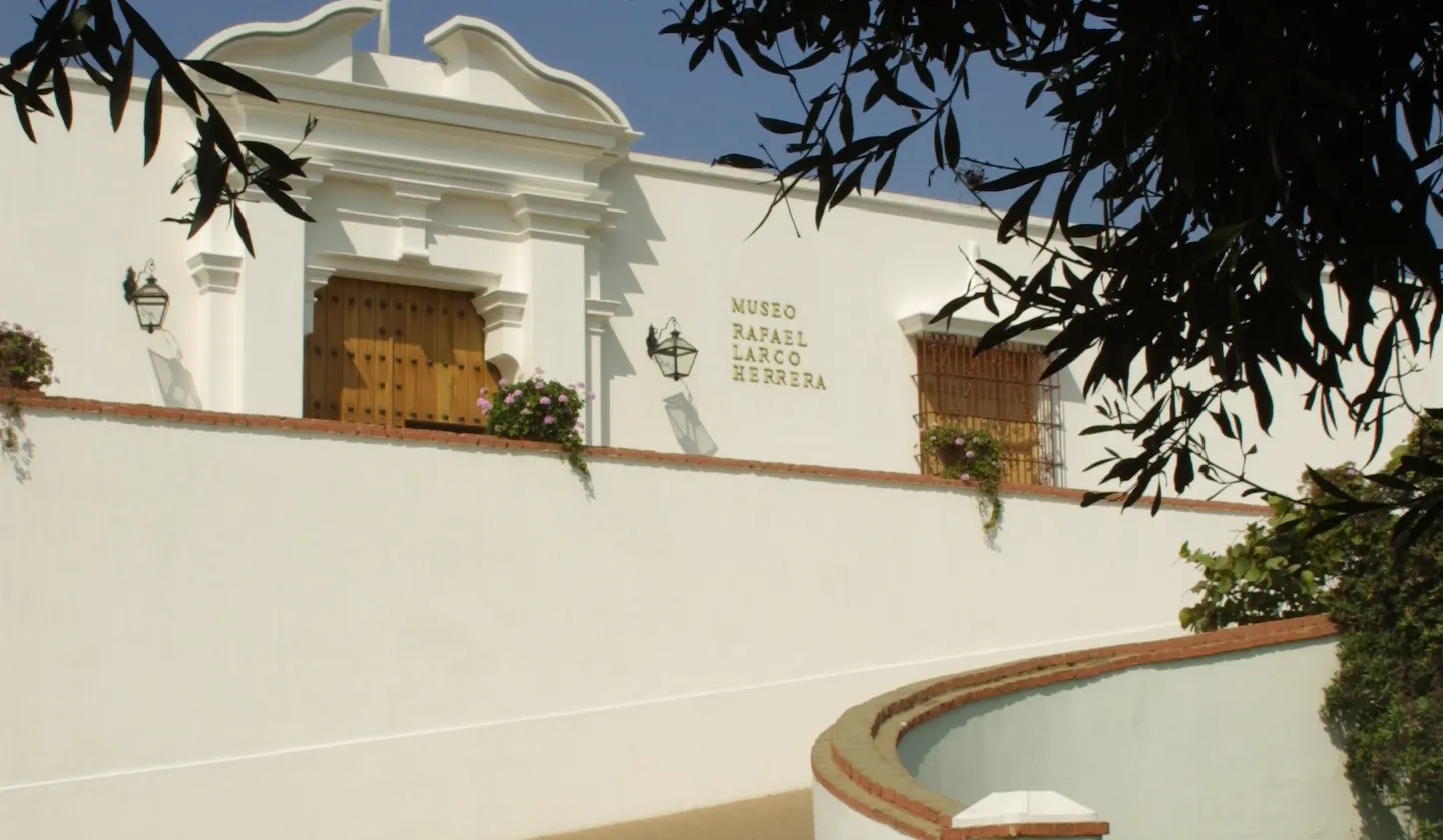 The whitewashed wall and carved wooden door of Museo Larco in Pueblo Libre, Lima, with the museum's name cut into the plaster and geraniums along the terrace below