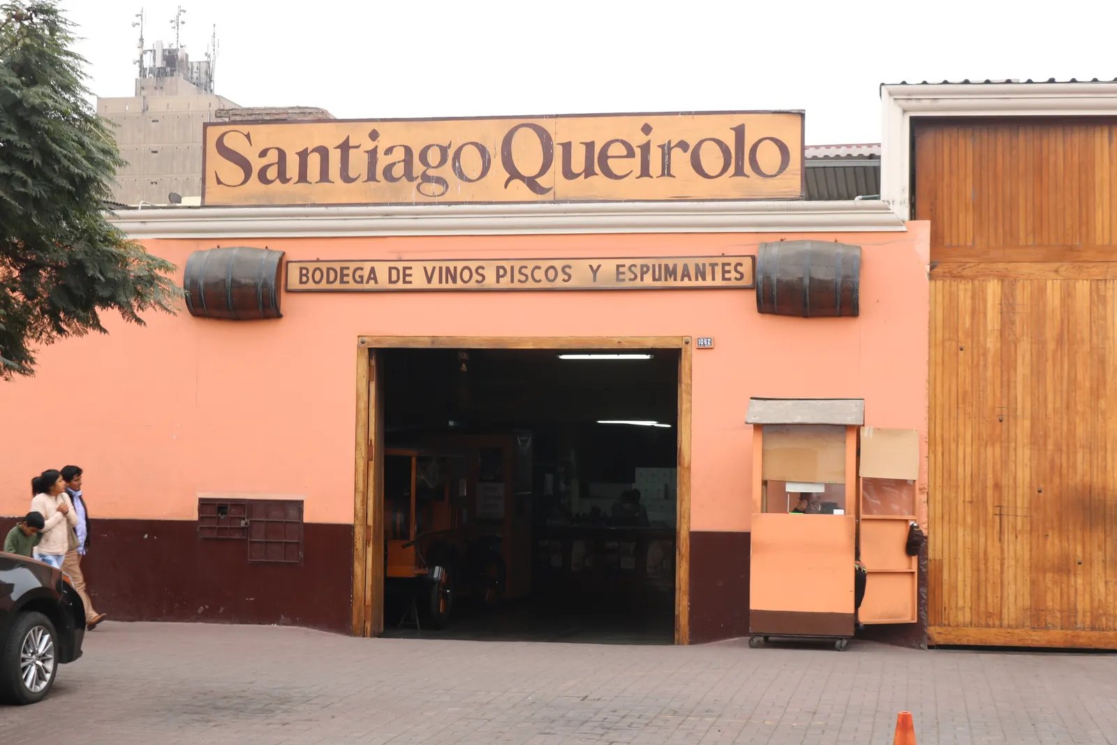 The salmon-pink front of the Antigua Taberna Queirolo in Pueblo Libre, Lima, with a painted sign over its open doorway and a wooden warehouse gate alongside