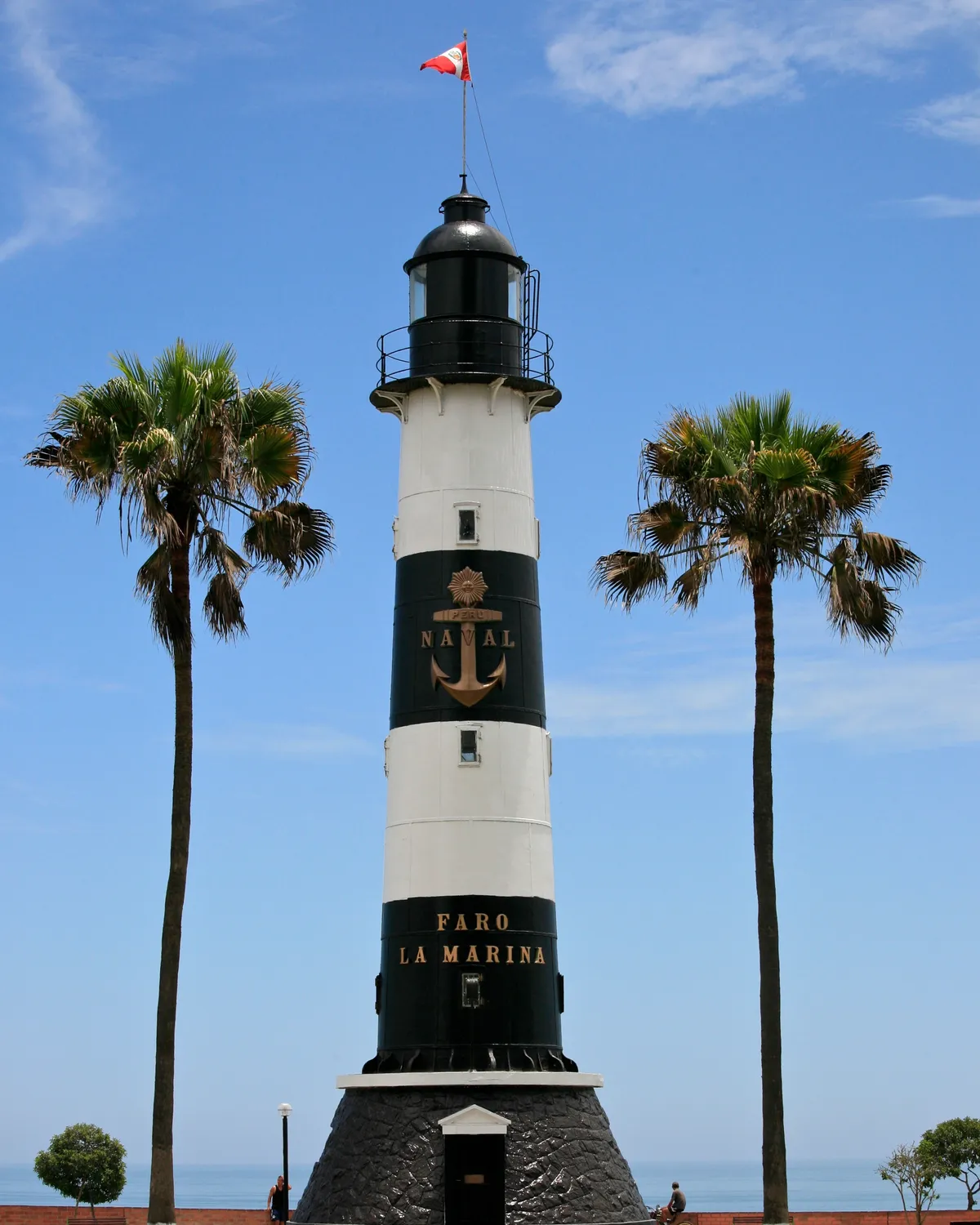 The black-and-white banded Faro La Marina lighthouse on the Miraflores clifftop, flying a Peruvian flag and flanked by two tall palms, with the Pacific behind it