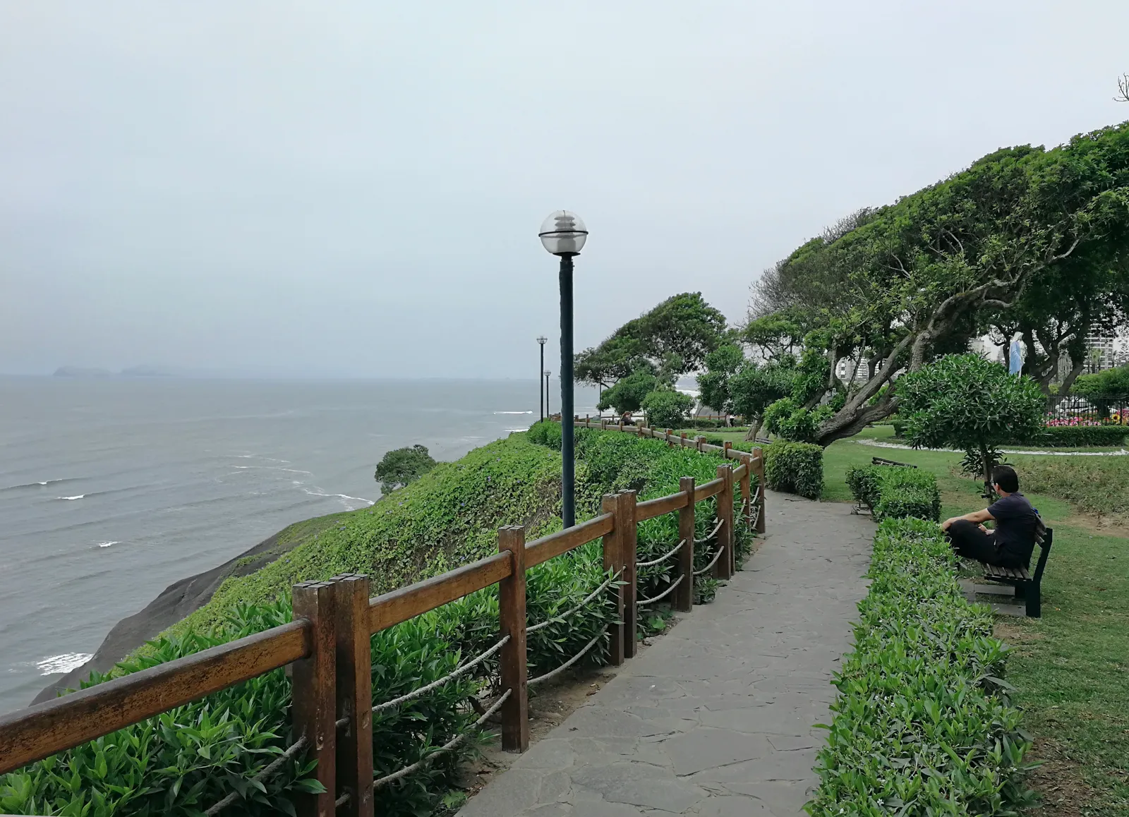 A paved clifftop path along the Malecón in Miraflores, fenced with wooden rails above the Pacific, with clipped hedges and a bench under wind-bent trees