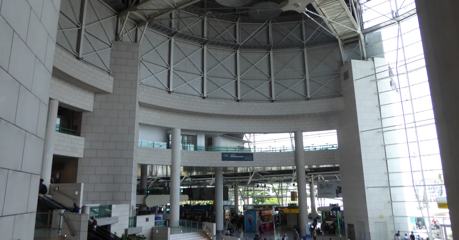The tall atrium of Terminal 1 at Lisbon's Humberto Delgado airport, with a steel-trussed roof, a curved upper gallery and travelers crossing the floor below