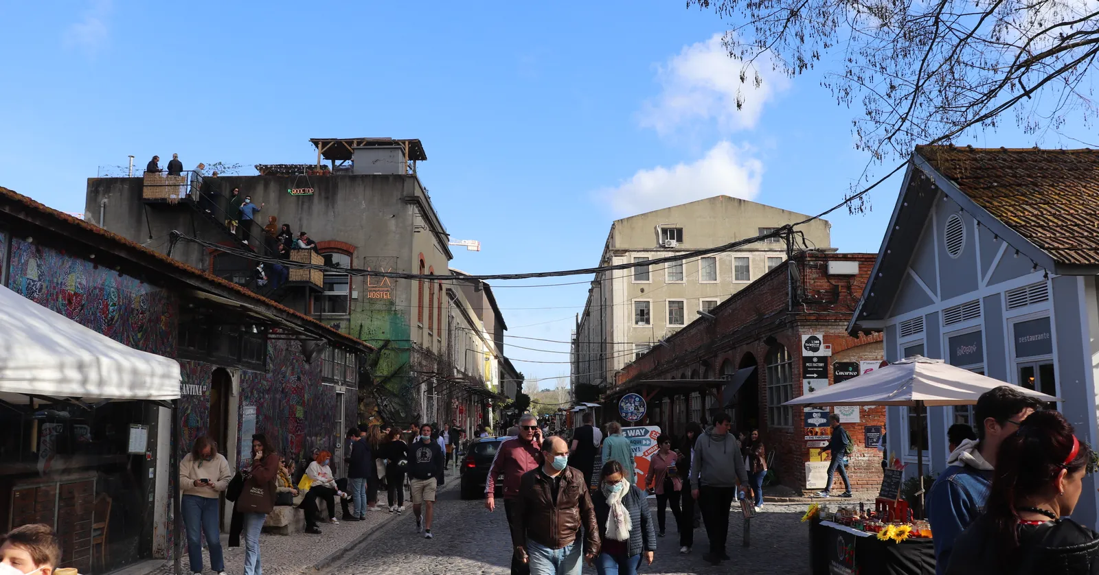 The main lane through LX Factory in Lisbon, lined with brick and stucco warehouse buildings, market awnings and people walking between them