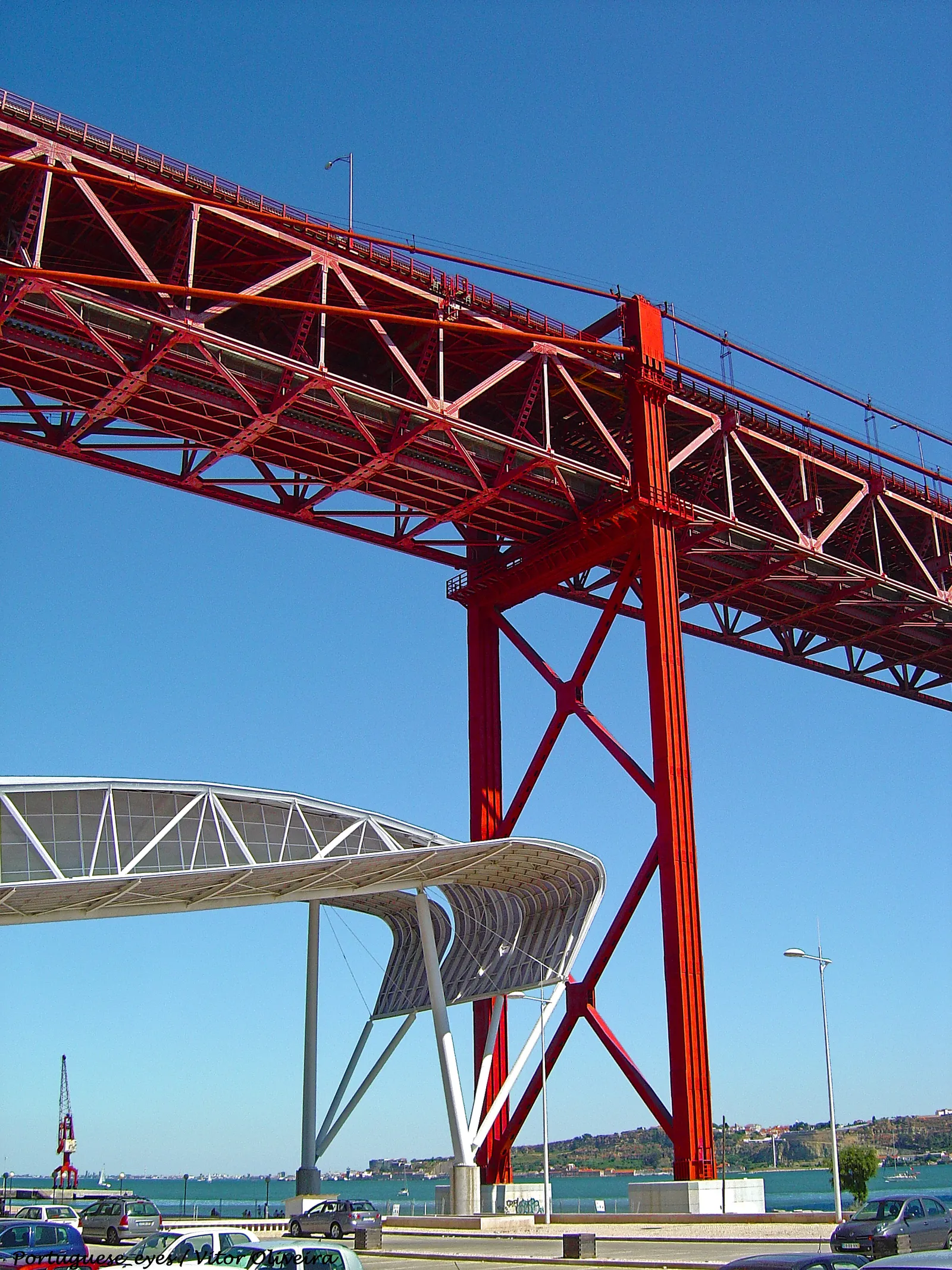 The red steel underside of the 25 de Abril bridge in Lisbon carried on a tall painted pier, seen from the ground in Alcântara