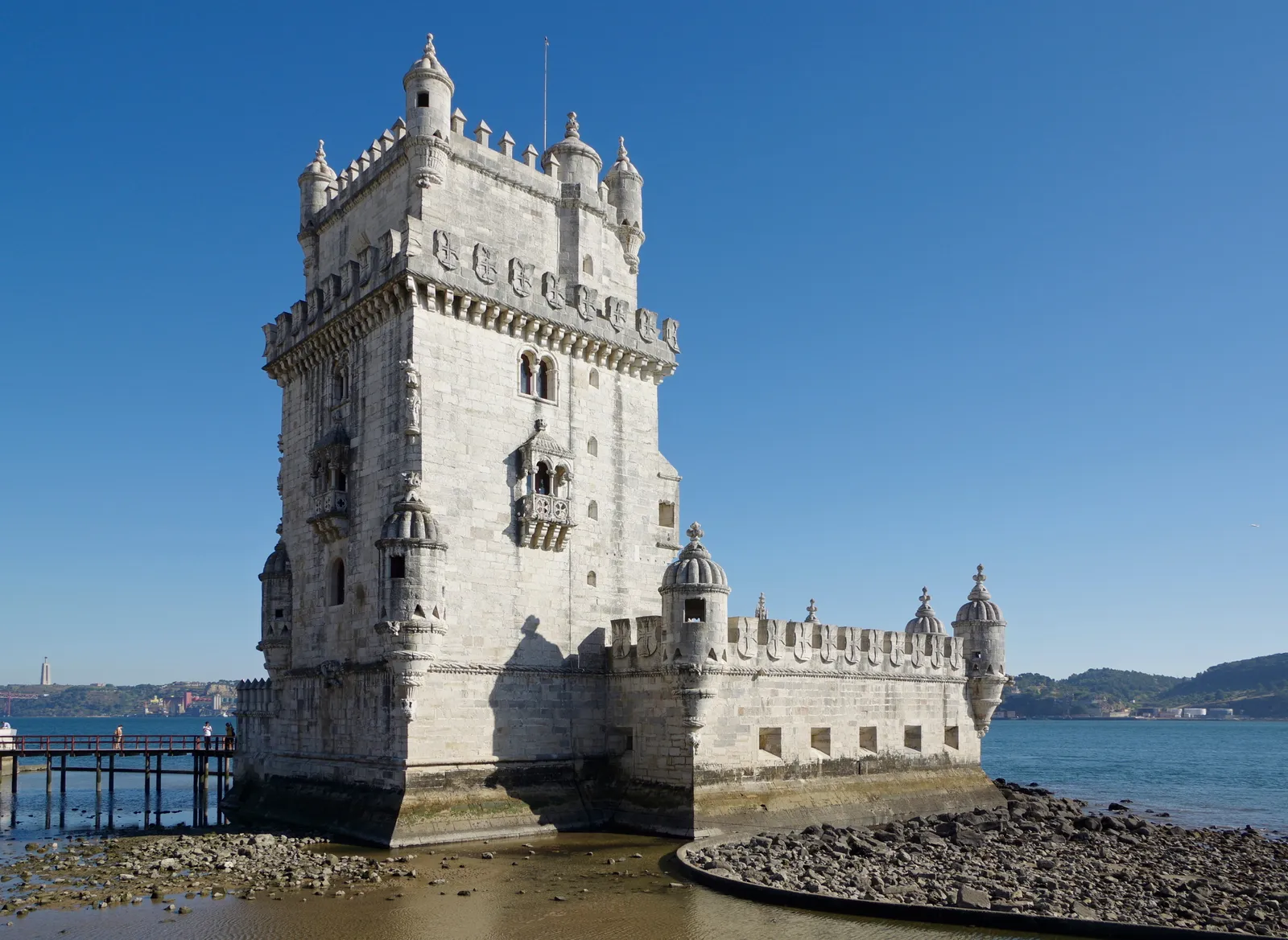 Belém Tower standing on the shoreline of the Tagus in Lisbon at low tide, its carved balconies and battlemented turrets against a blue sky