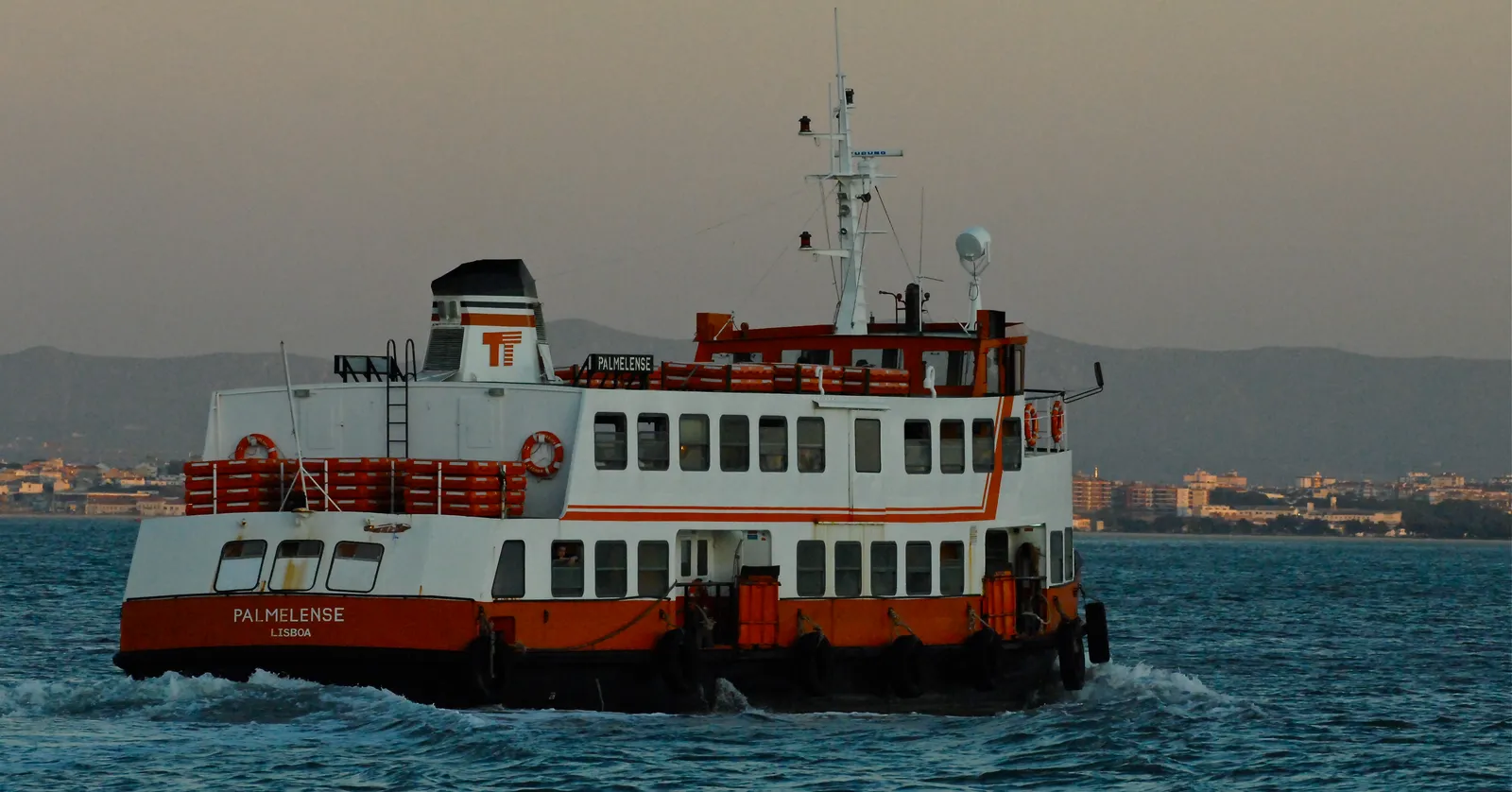 An orange and white Lisbon commuter ferry crossing the Tagus at low evening light, with the far shore behind it