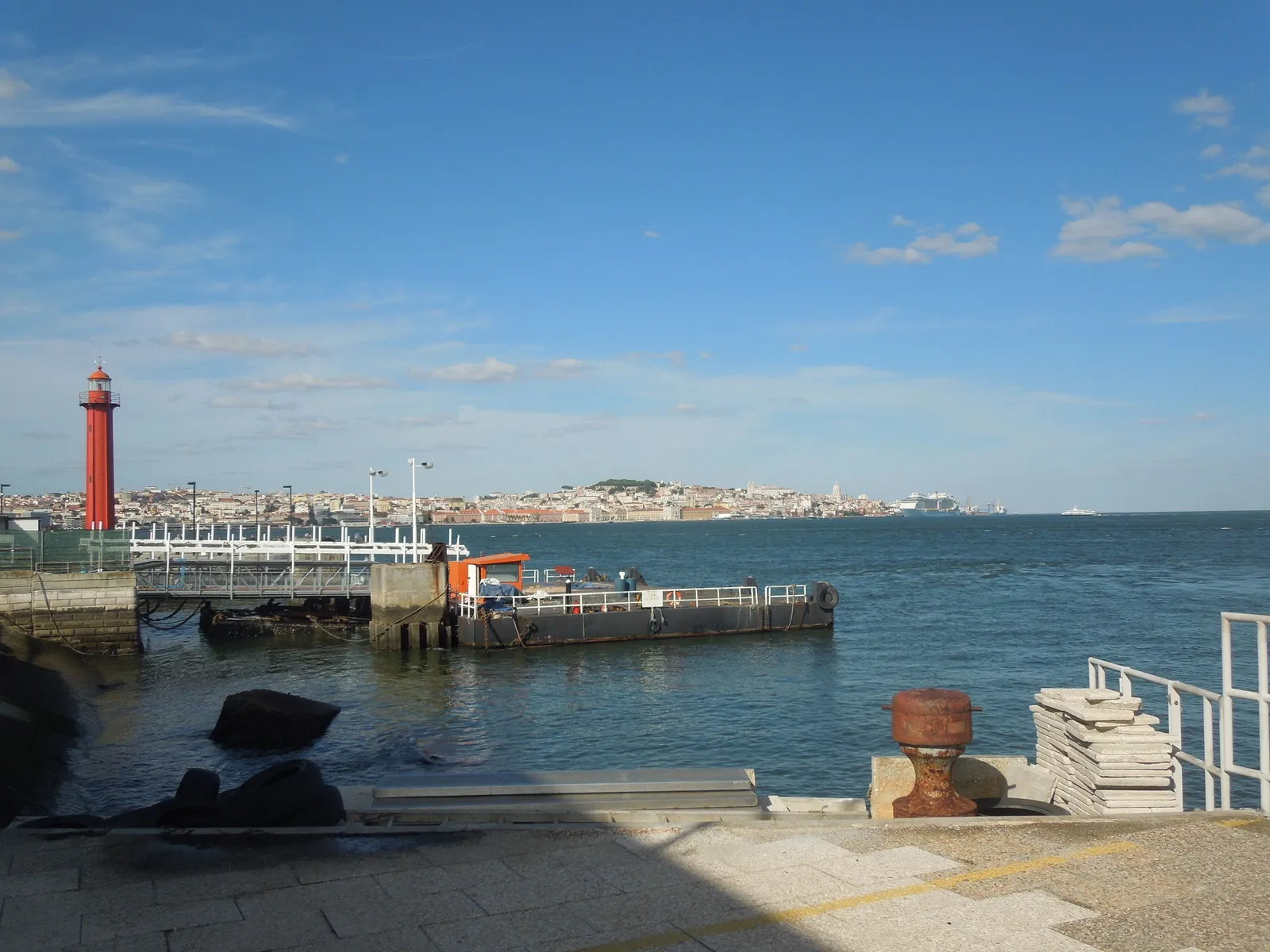 Lisbon seen across the Tagus from the quay at Cacilhas, with a red lighthouse and a jetty in the foreground and the city spread along the far bank