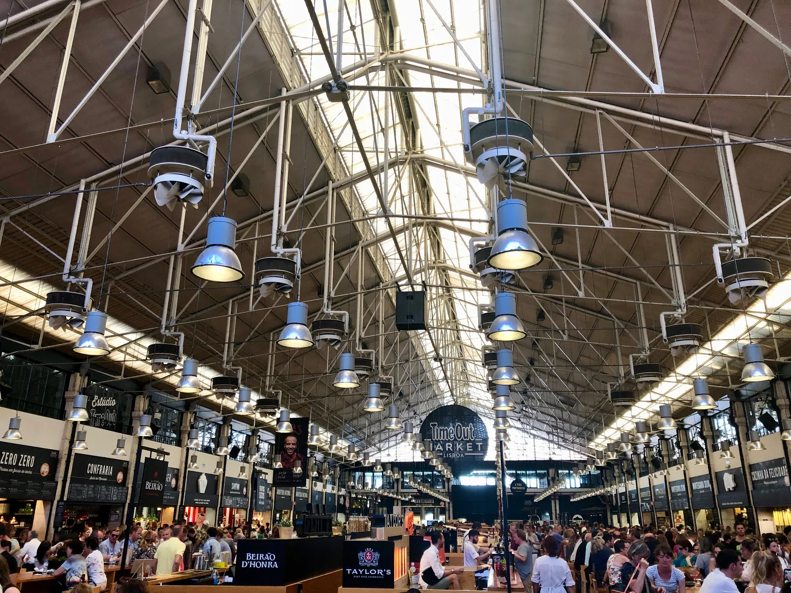 The steel roof and hanging lamps of the Mercado da Ribeira food hall in Lisbon, with crowded communal tables below and food counters along both sides