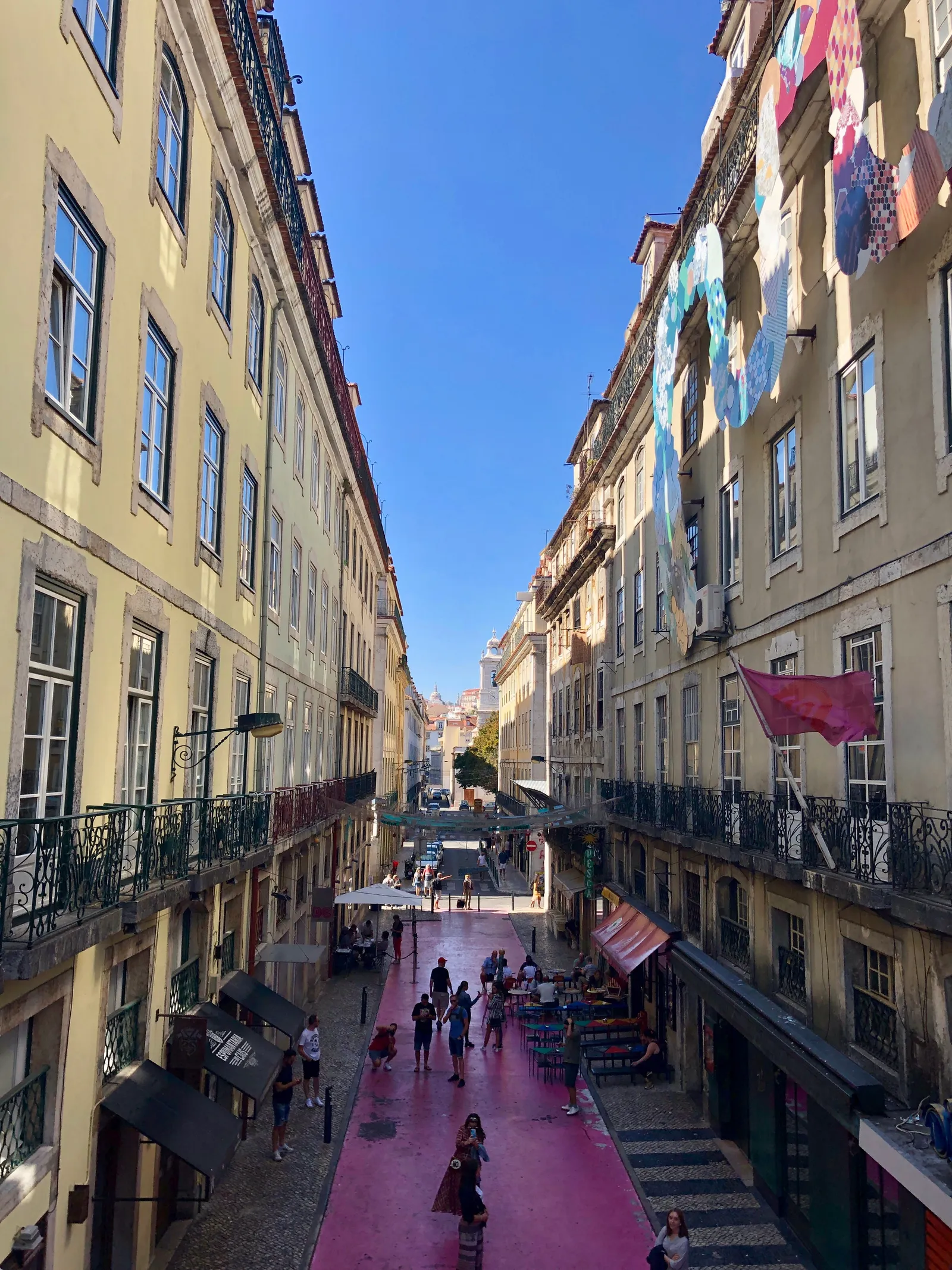 Rua Nova do Carvalho in Lisbon seen from above, its roadway painted bright pink between yellow buildings with bar terraces along both sides