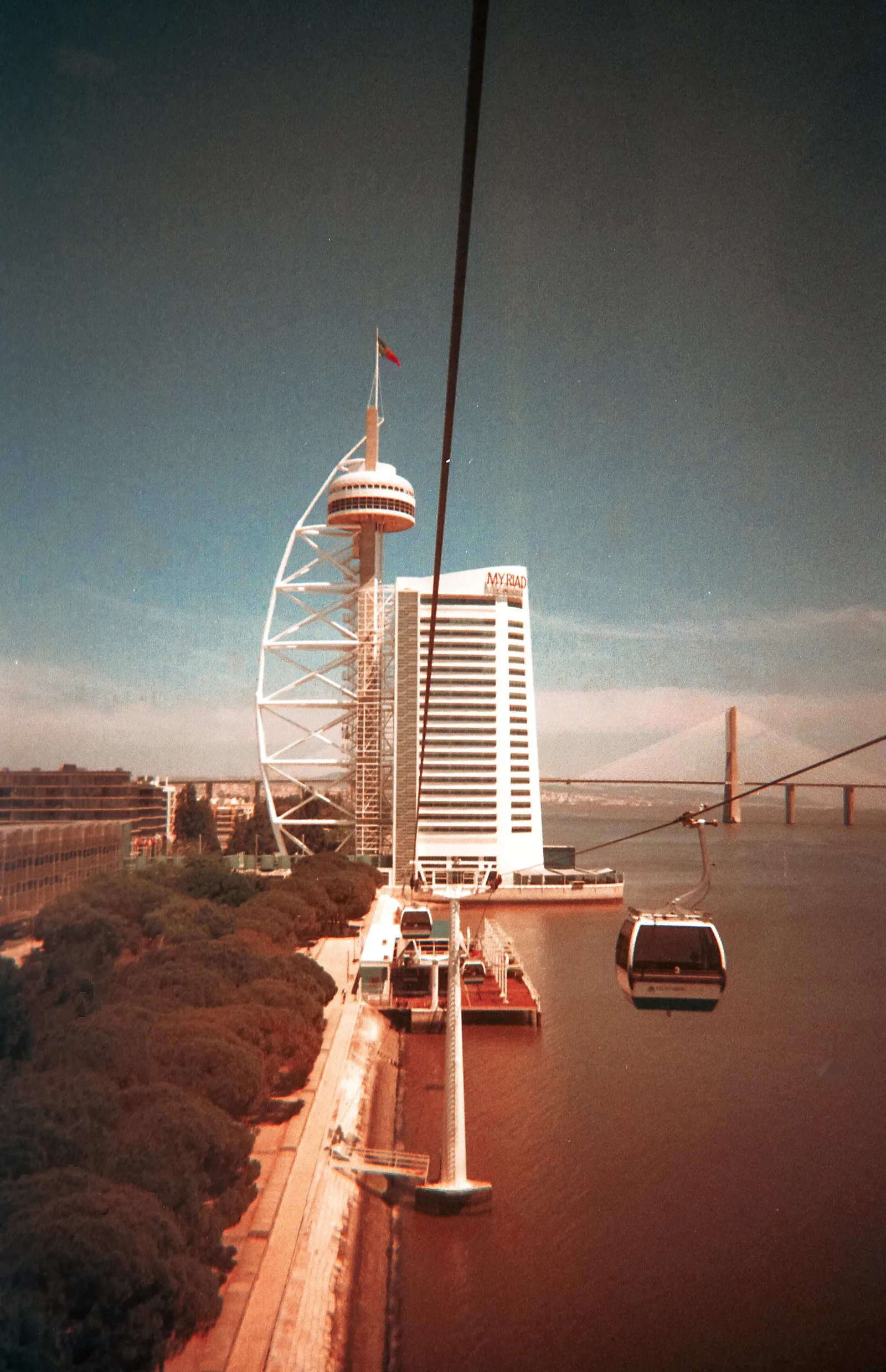 A cable car cabin crossing the Tagus waterfront at Parque das Nações in Lisbon, with the sail-shaped Vasco da Gama tower ahead and the Vasco da Gama bridge in the distance