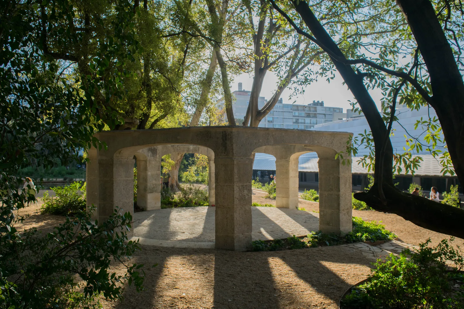 A stone colonnade standing among trees in the garden of the Gulbenkian modern art centre in Lisbon, with the white roof of the museum building behind