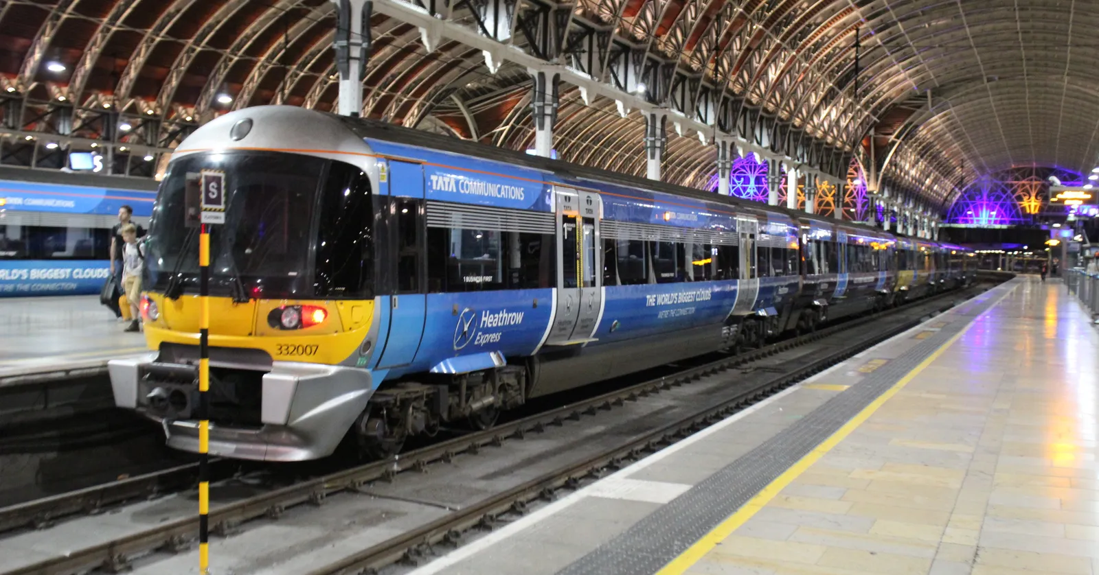 A blue and yellow Heathrow Express train at a platform under the arched iron and glass roof of London Paddington station, lit up in the evening