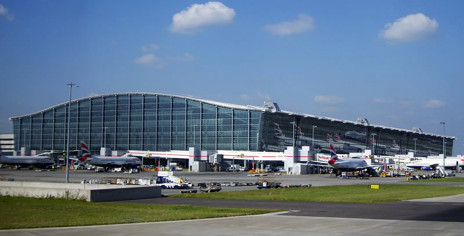 The long glass and steel front of Heathrow's Terminal 5 under a blue sky, with British Airways planes parked at the stands in front of it
