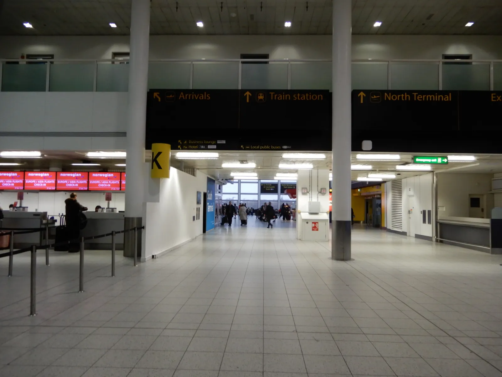 The concourse of Gatwick Airport's South Terminal, with overhead signs pointing to arrivals, the train station and the North Terminal, and check-in desks to one side