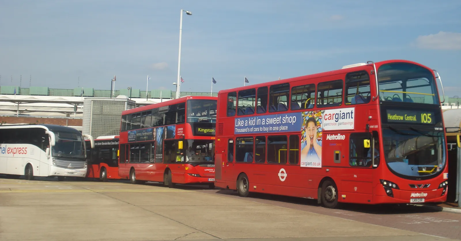 A white long-distance coach and red double-decker buses lined up at Heathrow's Central Bus Station between Terminals 2 and 3, under a blue sky