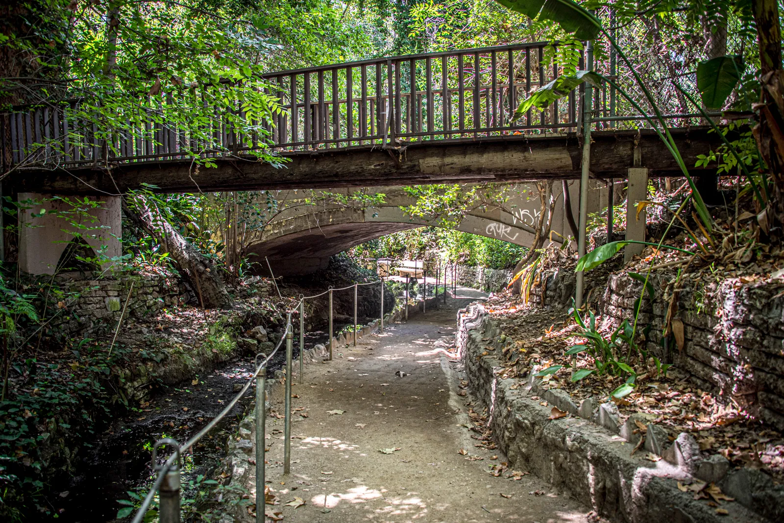The path through Fern Dell in Griffith Park, a shaded gravel walkway beside a stone-lined stream under heavy tree cover, with an old wooden footbridge crossing overhead