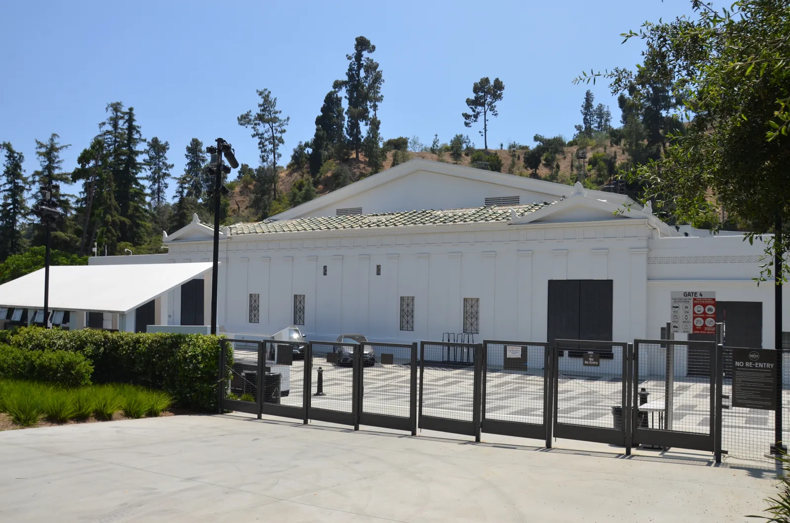 The Greek Theatre in Griffith Park, a low white building with a columned classical facade and a tiled roof, set against a wooded hillside behind steel gates