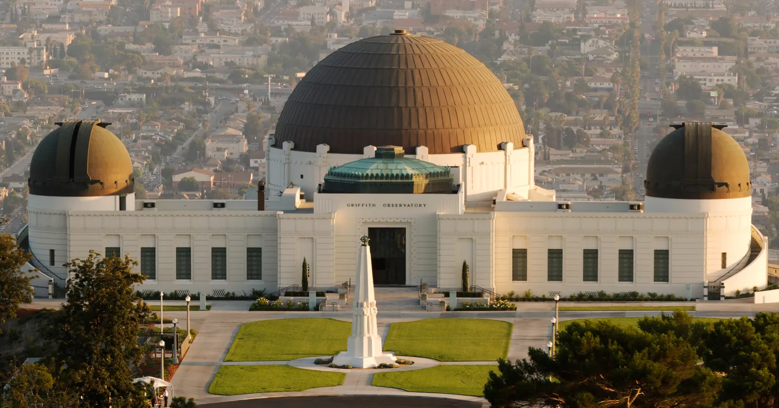 Griffith Observatory in Los Angeles seen from the front lawn in late sun, a white art deco building with a large central copper dome and two smaller domes either side, a tall obelisk on the grass in front and the city spreading out behind