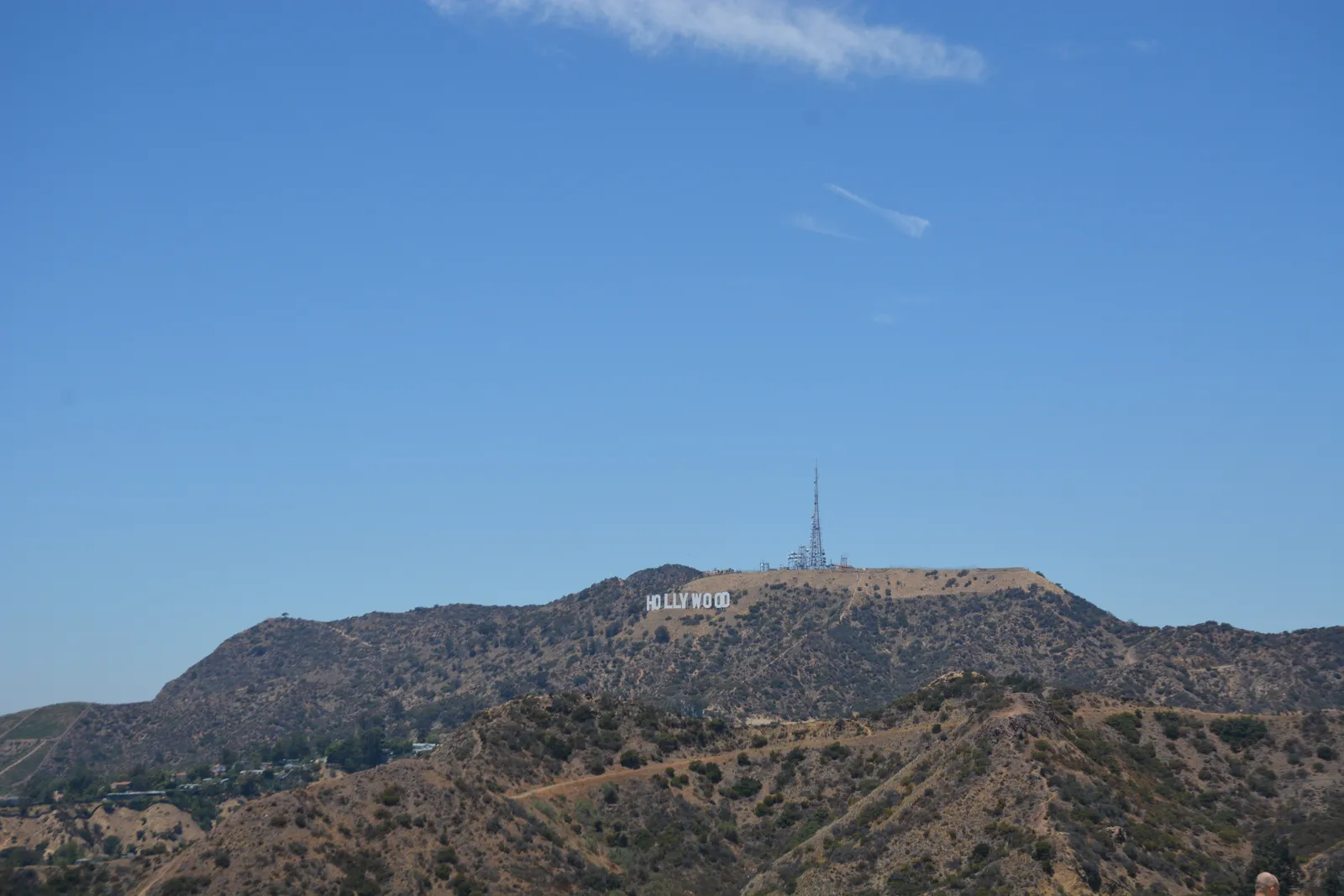 The Hollywood Sign seen from Griffith Observatory, white letters small and level on the brown ridge of Mount Lee with a broadcast mast above them and dry scrub hillside in the foreground
