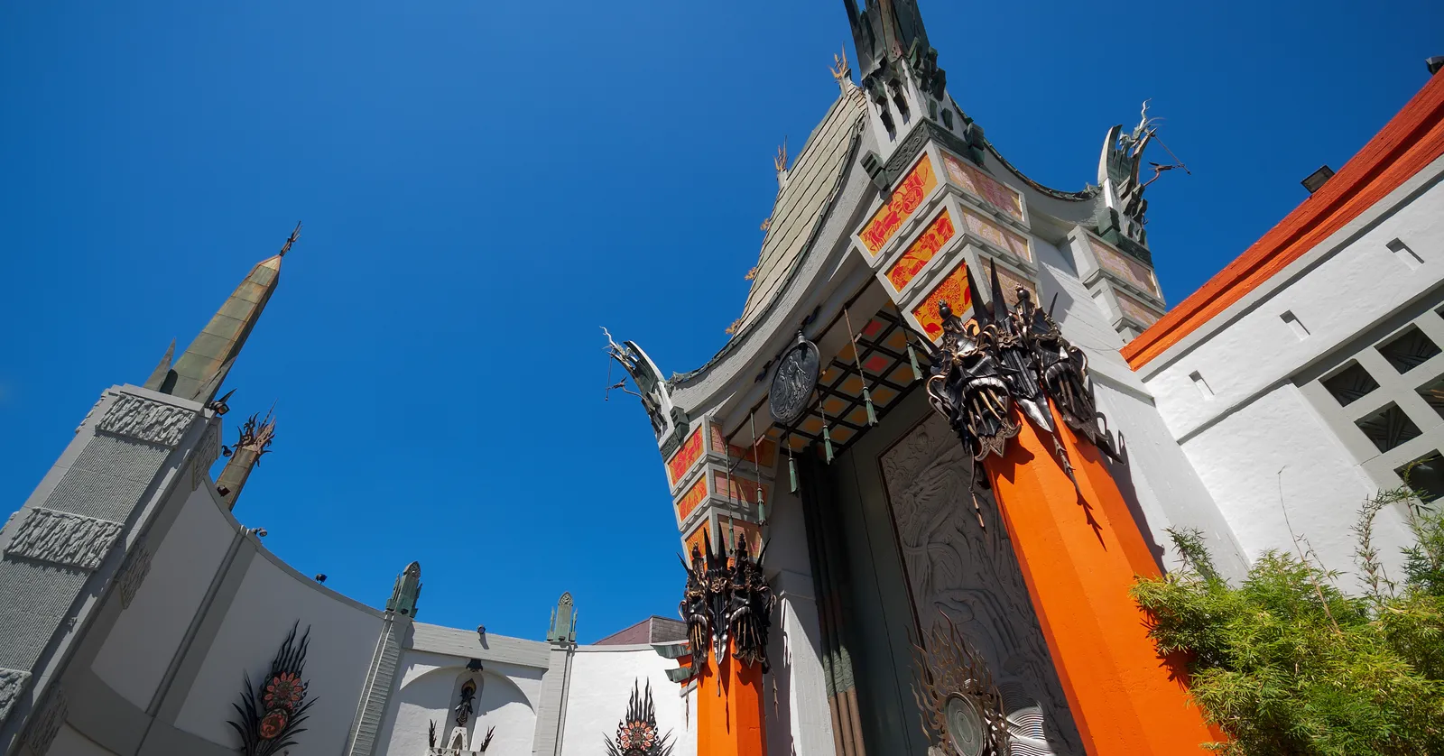 The TCL Chinese Theatre on Hollywood Boulevard seen from below, its green pagoda roof and bronze finials rising above red columns and a carved stone entrance, against a cloudless blue sky