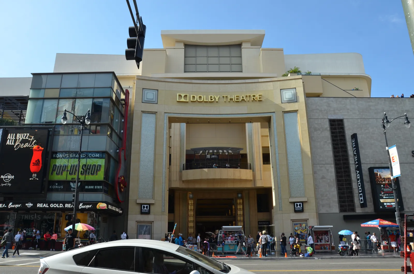The Dolby Theatre entrance on Hollywood Boulevard, a pale stone arch with the theatre's name in gold lettering across the top, shops either side and people crossing the pavement in front of it