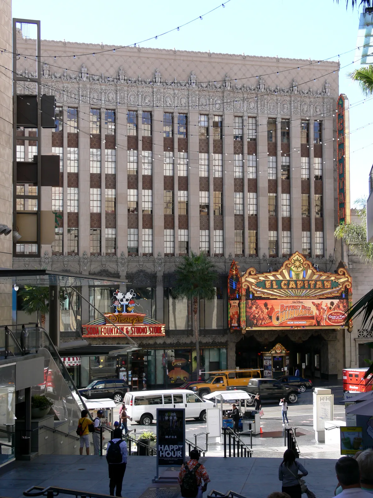 The El Capitan Theatre on Hollywood Boulevard, a patterned grey stone office block above a gold and red painted marquee lit with bulbs, with traffic and pedestrians on the street below