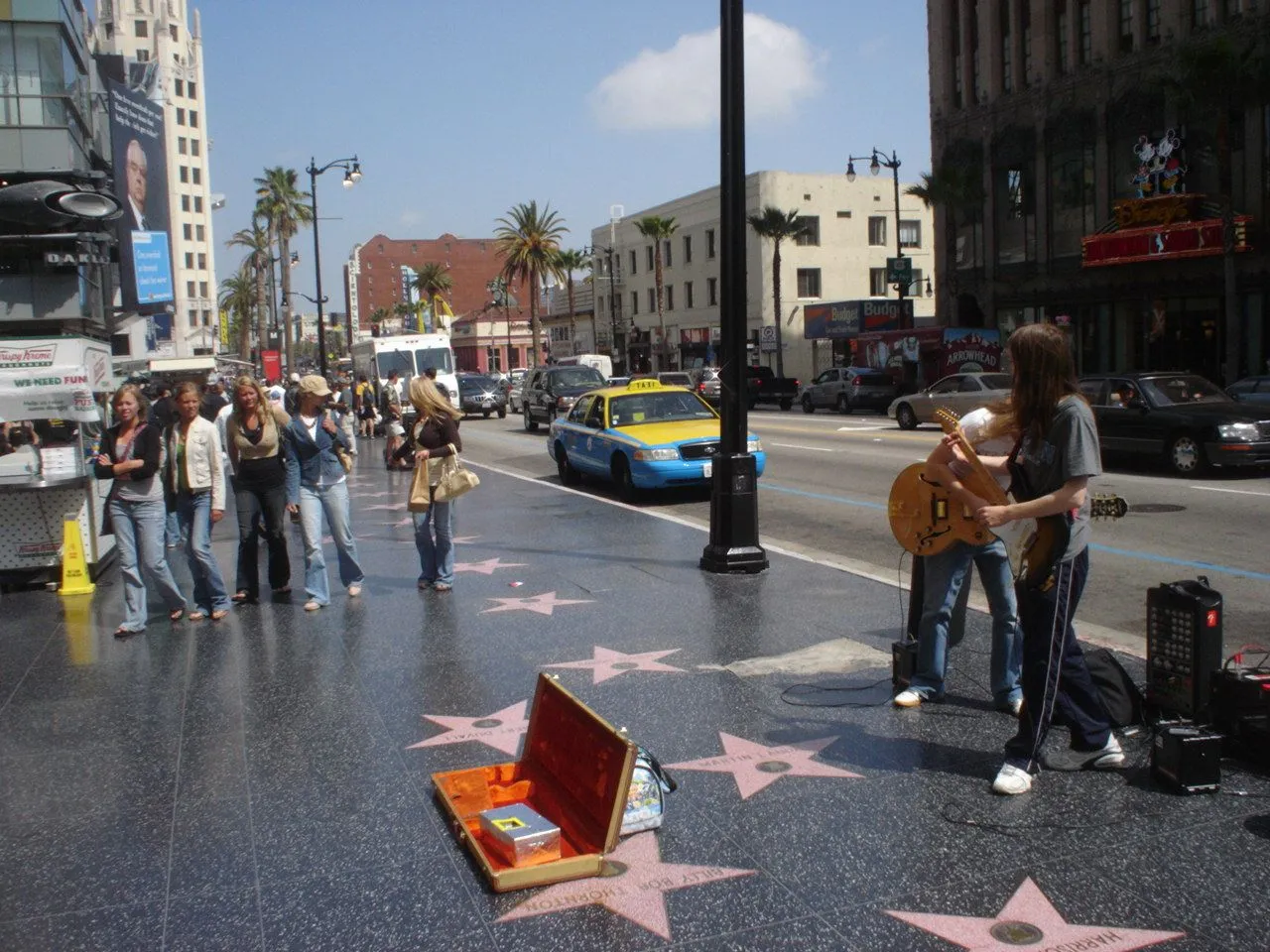 Pink terrazzo stars set into the wide pavement of Hollywood Boulevard, with a busker playing guitar beside an open guitar case, pedestrians walking past and traffic on the road behind