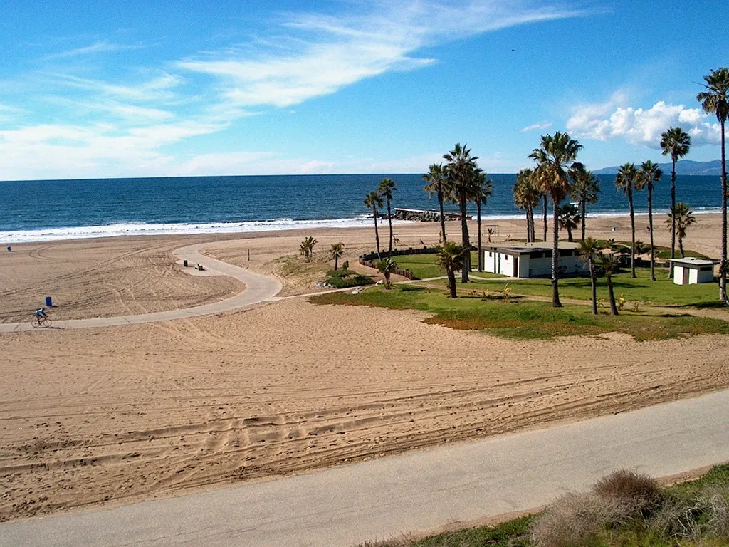 Dockweiler State Beach in Los Angeles, a wide flat expanse of pale sand with a paved path curving through it, a row of palm trees and low white buildings on the grass, and the Pacific beyond