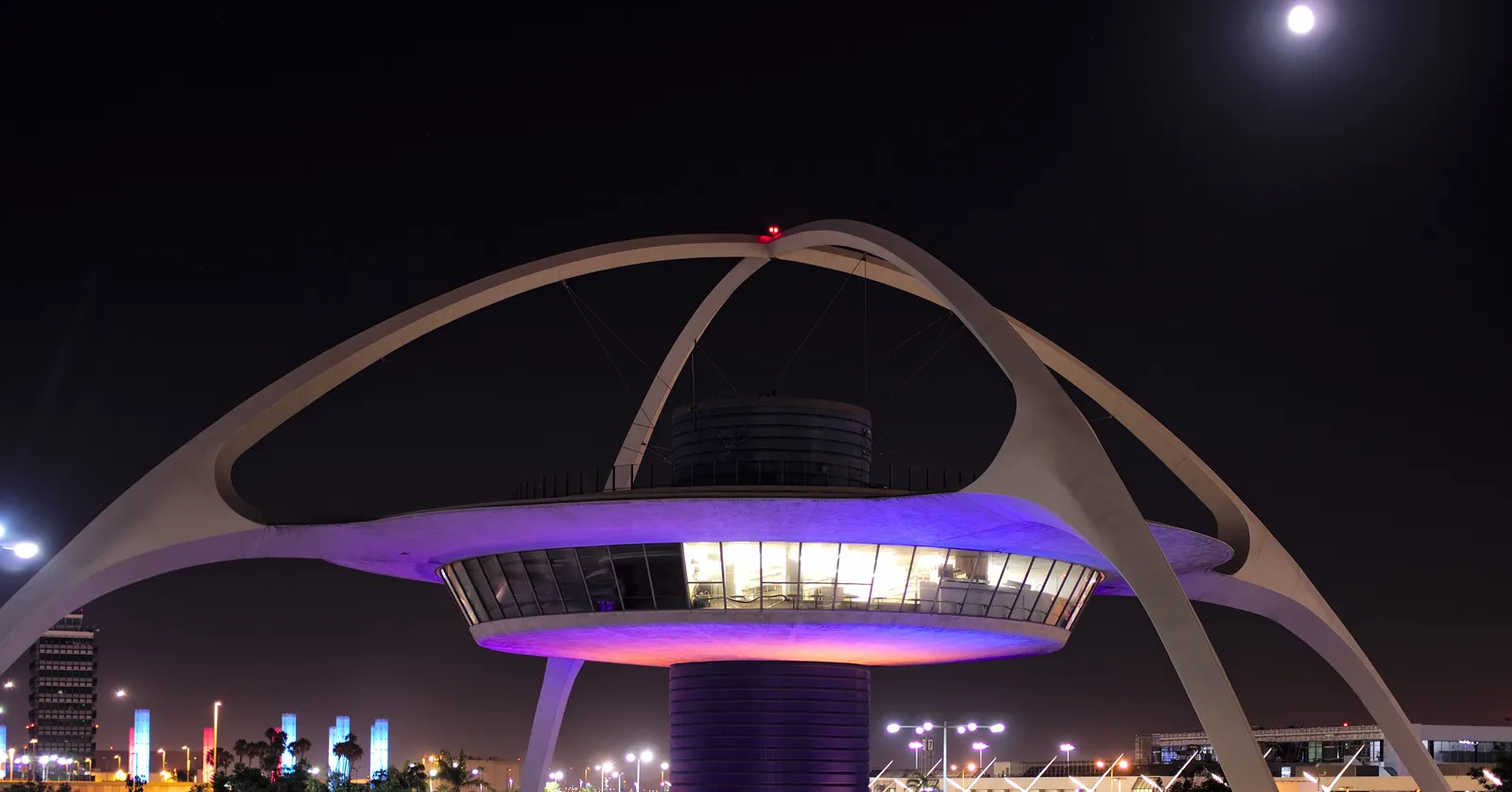 The Theme Building at Los Angeles International Airport lit purple after dark, its white parabolic arches crossing above a disc-shaped glass restaurant level on a central column, with a full moon in the black sky beside it