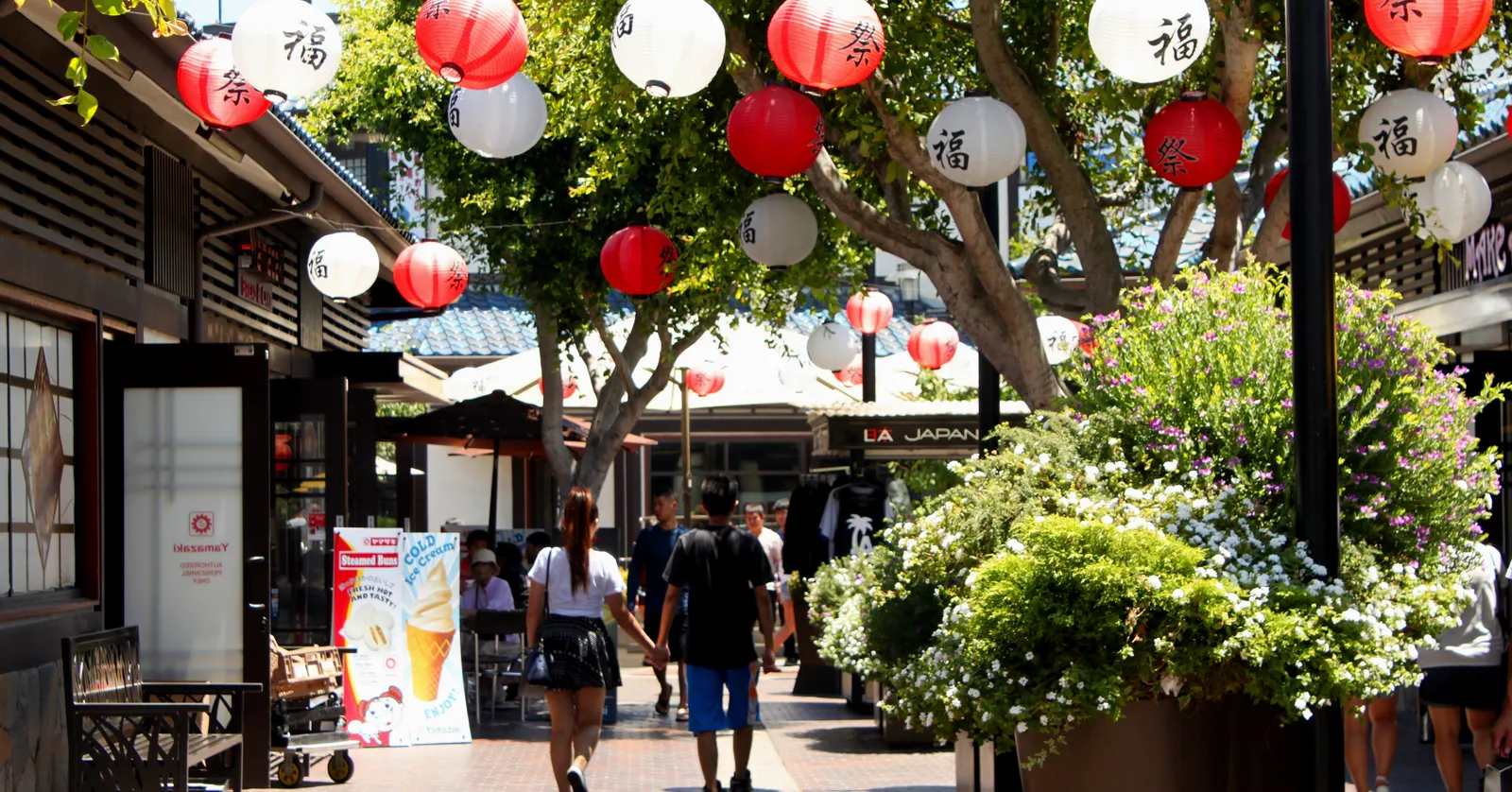 A pedestrian lane in Japanese Village Plaza in Los Angeles, red and white paper lanterns strung between trees overhead, low tiled shopfronts down both sides and people walking the brick paving