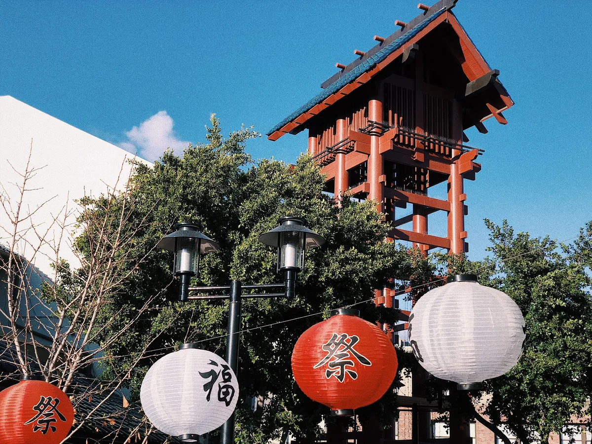 The wooden fire tower at Japanese Village Plaza in Los Angeles, a tall red timber frame carrying a small blue-tiled roof, rising above trees with white and red paper lanterns strung in front of it