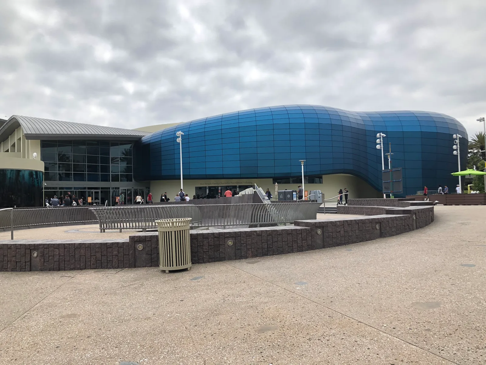 The Aquarium of the Pacific in Long Beach, a low building clad in curved deep-blue glass panels shaped like a breaking wave, with visitors queuing at the entrance under a grey sky