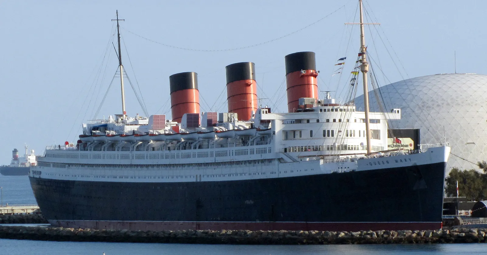 The RMS Queen Mary moored at Long Beach, a long black-hulled ocean liner with a white superstructure and three red funnels banded in black, with a white geodesic dome behind her stern