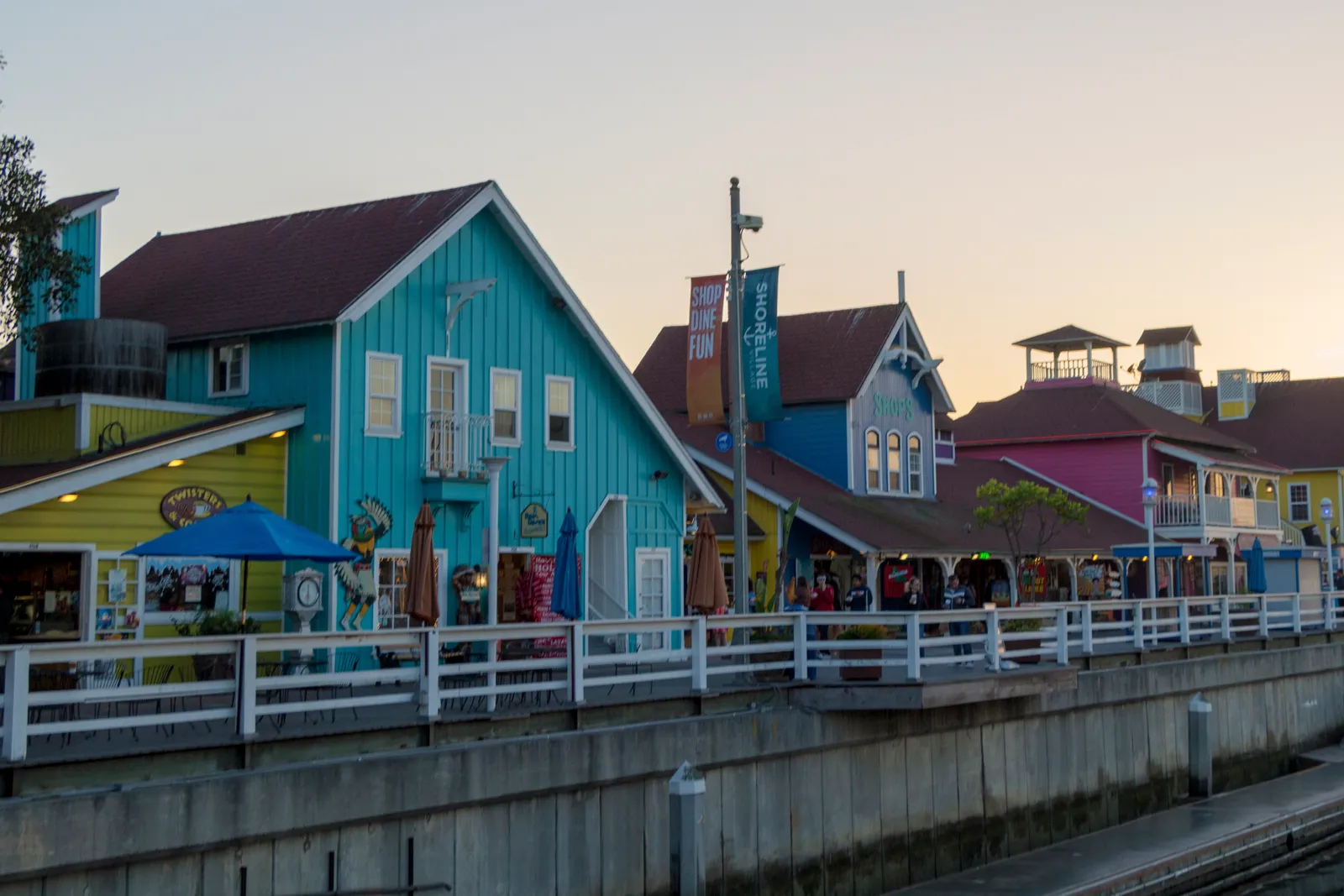 Shoreline Village in Long Beach at dusk, a row of brightly painted turquoise, blue and pink clapboard buildings with steep gables, fronted by a white-railed boardwalk above the harbour water