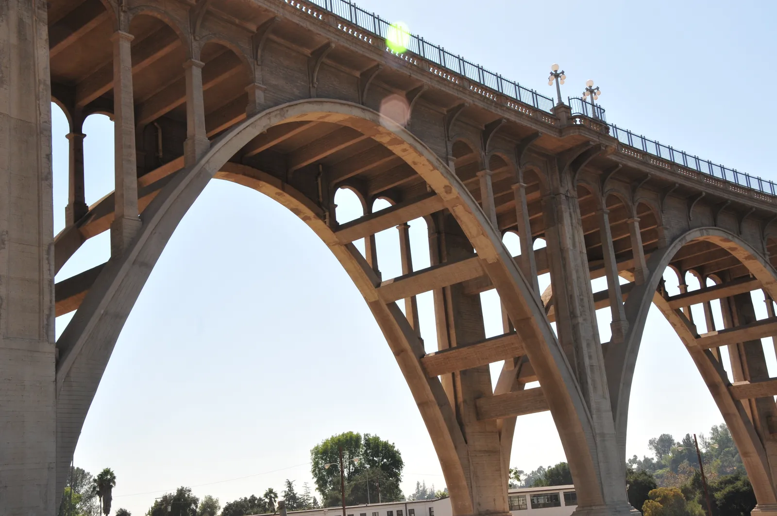 The Colorado Street Bridge in Pasadena seen from beneath, tall concrete arches carrying a roadway with a balustrade and lamp posts along its edge, high above the trees of the Arroyo Seco