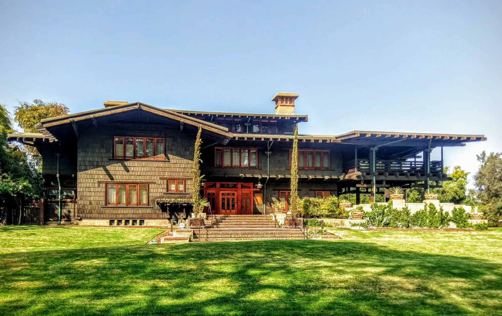 The Gamble House in Pasadena, a broad two-storey timber Craftsman house with deep overhanging eaves, exposed rafter ends and a wide terrace, standing on a lawn under a clear sky