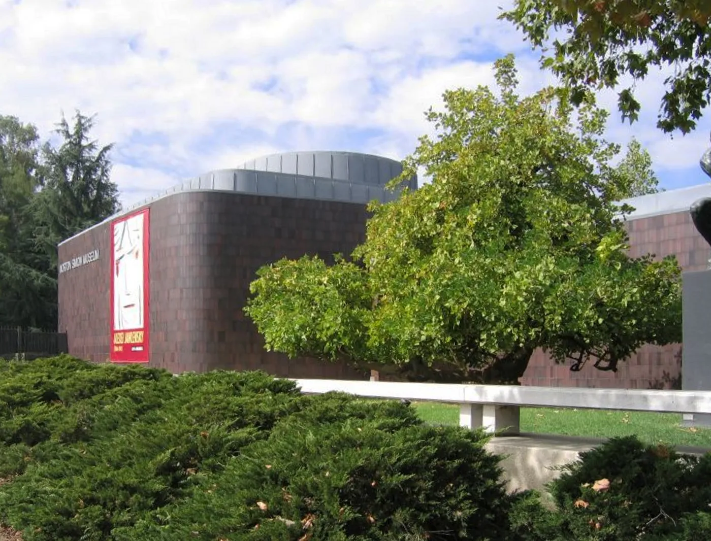 The Norton Simon Museum in Pasadena, a low curved building faced in dark brown tile with a banner beside its entrance, set back behind mature trees and clipped hedging
