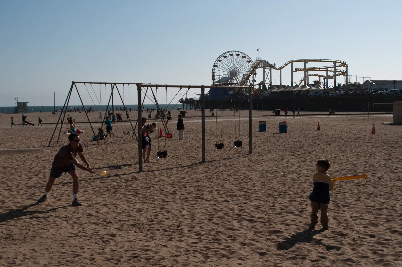The open-air gymnastics equipment at Muscle Beach in Santa Monica, steel frames hung with rings standing directly on the sand, with the pier's Ferris wheel and roller coaster behind