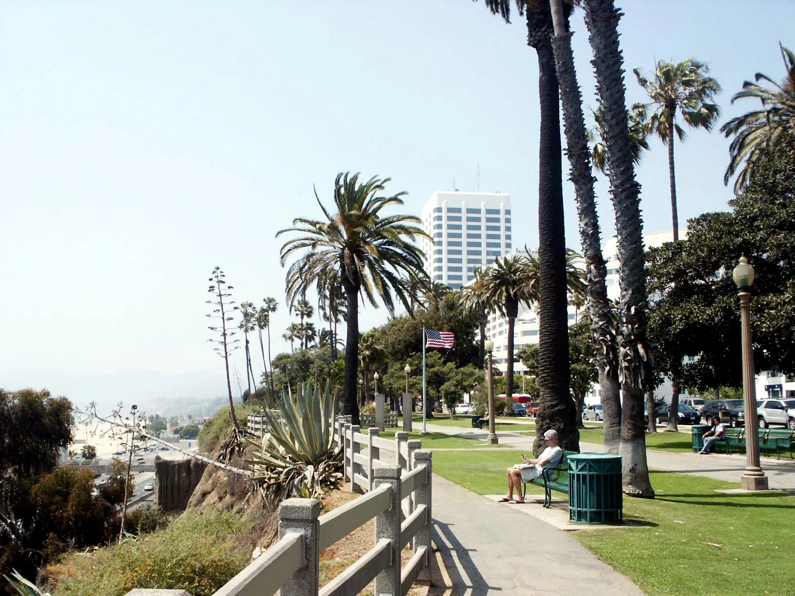 The path through Palisades Park in Santa Monica, running along the top of the coastal bluff behind a white fence, with palm trees, benches and the shoreline dropping away to the left