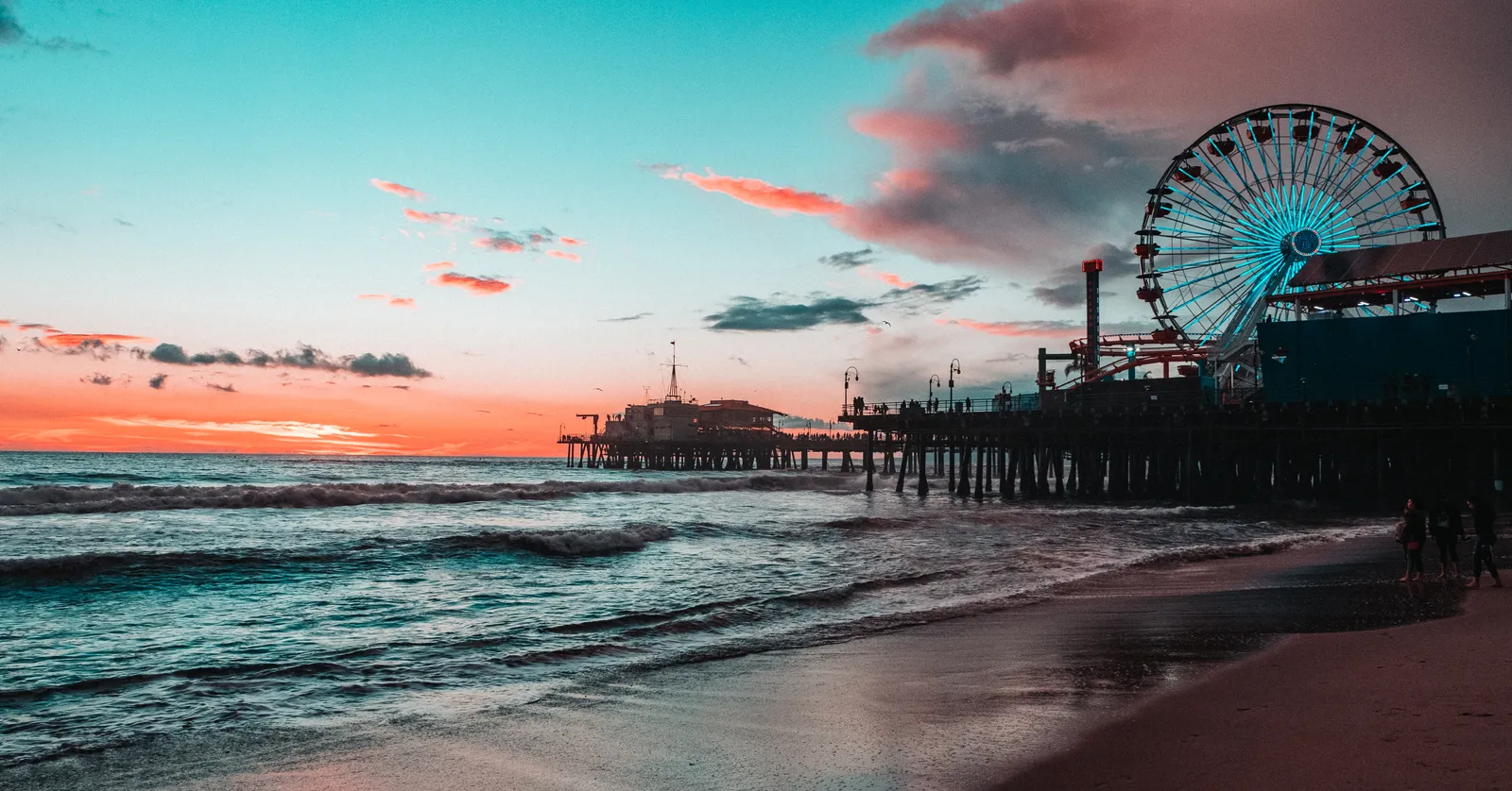Santa Monica Pier at sunset seen along the beach, the lit Ferris wheel and amusement rides standing on the pier deck above the pilings, with surf running up the sand in the foreground