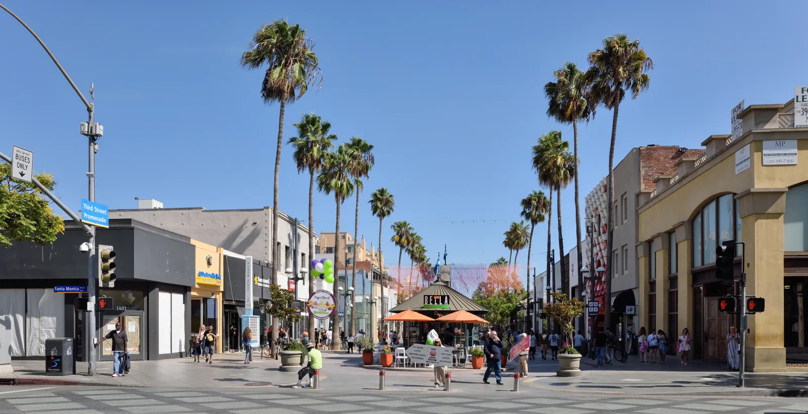 Third Street Promenade in Santa Monica, a wide pedestrian street with no cars on it, lined with shopfronts and tall palm trees, with people walking down the middle under a clear sky