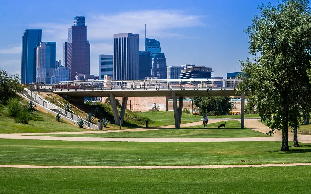 Los Angeles State Historic Park, a wide mown lawn and gravel path with a long concrete pedestrian bridge crossing the middle distance and the downtown Los Angeles towers standing on the horizon behind it