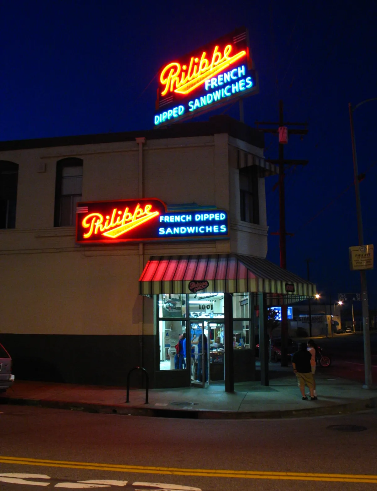 The corner entrance of Philippe the Original in Los Angeles after dark, two lit neon signs reading French Dipped Sandwiches on the cream-painted building above a striped awning, with customers visible through the windows