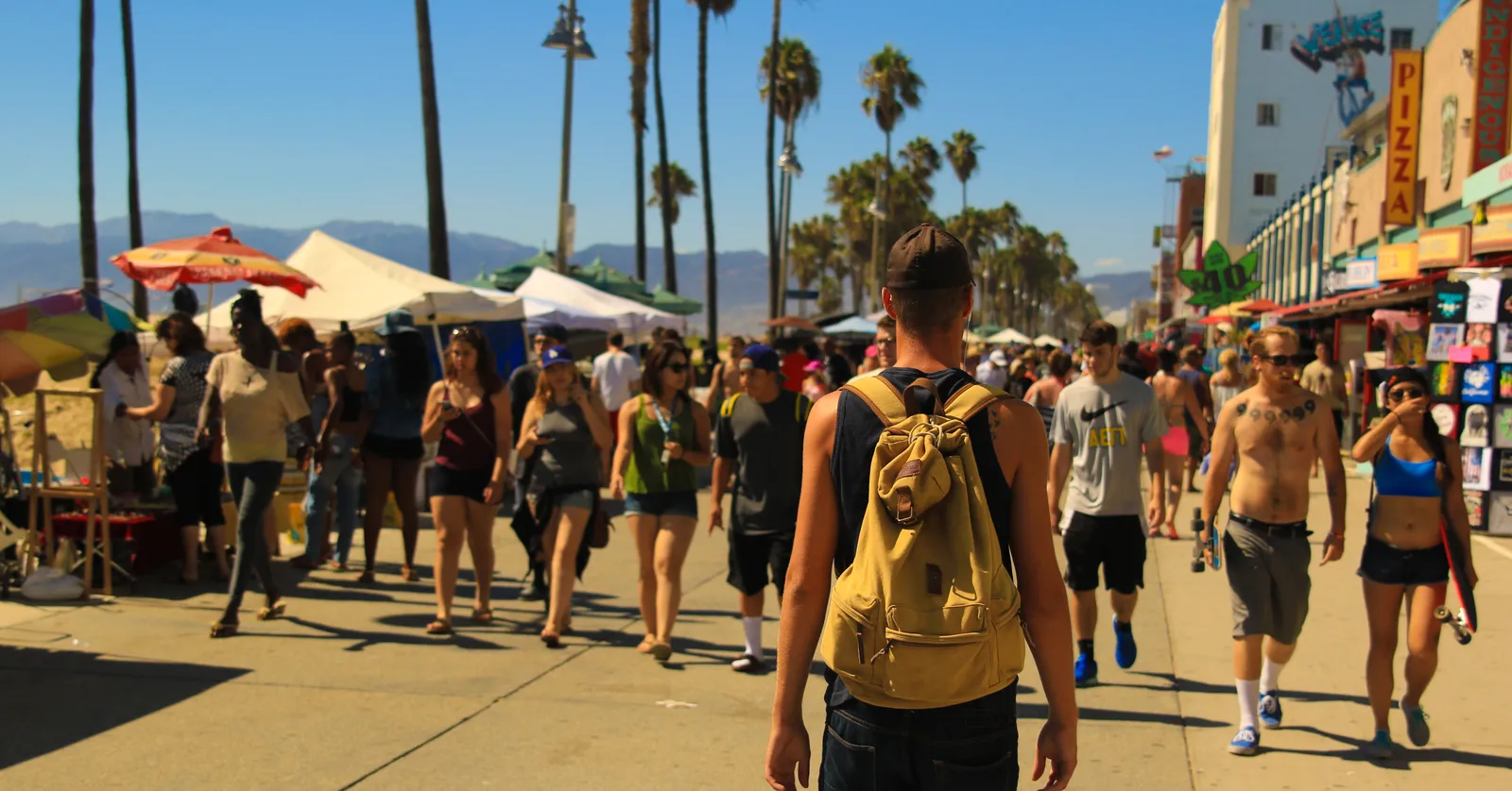 The crowded Ocean Front Walk at Venice Beach in Los Angeles, a wide paved promenade packed with people walking between market stalls and low shopfronts, with tall palms down the middle and hills in the distance