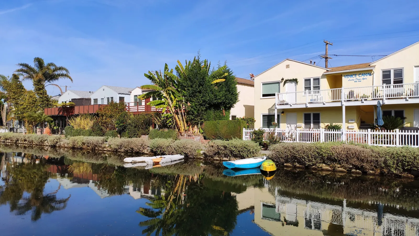 One of the Venice canals in Los Angeles, a narrow strip of still water reflecting the houses along both banks, with small rowing boats moored at the edge, palms and a clear blue sky