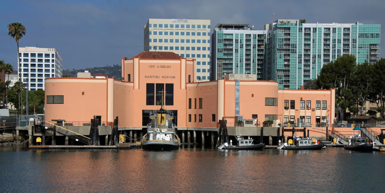 The pale pink Streamline Moderne ferry building that houses the Los Angeles Maritime Museum in San Pedro, seen across the water with small boats moored in front